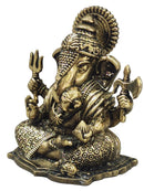 A brass Lord Ganesha idol with intricate carvings, seated on a lotus base and holding a trident and axe, photographed against a neutral gray background.