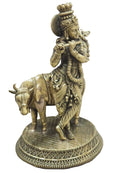 An ornate brass statue of Lord Krishna playing the flute, mounted on a decorated circular base with a cow at his side, set against a light gray background.